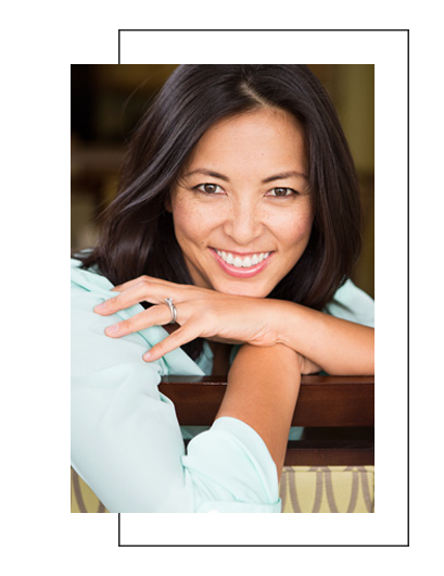 The image features a woman smiling at the camera with her hand on her chin, leaning slightly towards the viewer. She has dark hair, appears to be wearing makeup, and is dressed in a light-colored top.