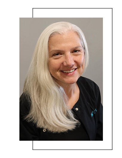 The image shows a smiling woman with long gray hair, wearing glasses and a black top, posing for a portrait headshot.