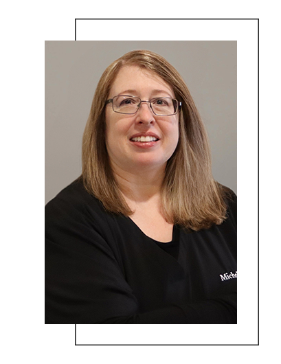 The image shows a woman with short hair wearing glasses, smiling at the camera. She has a light-colored top and appears to be seated against a neutral background.