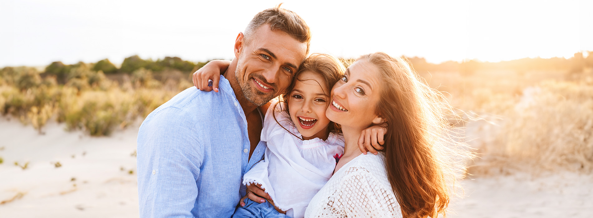 The image shows a family of four posing together on a sandy beach during sunset, with a clear sky and warm lighting.