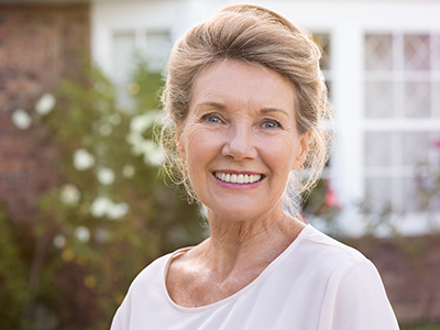 A smiling woman with short blonde hair, wearing a white top, stands in front of a house with a brick wall and a white door.