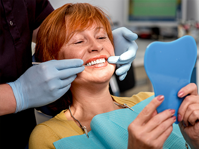 Woman sitting in dental chair with blue mouthguard, smiling at camera while holding toothbrush, being attended by dentist who is cleaning her teeth.