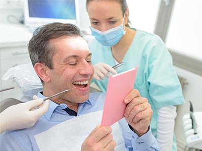 The image shows a man sitting in a dental chair, holding up a pink card with a surprised expression, while a female dental professional looks on with a smile, both in a dental office setting.