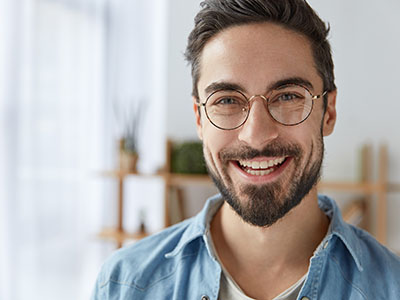 The image shows a man with a beard smiling at the camera. He has glasses, a goatee, and is wearing a light blue shirt.