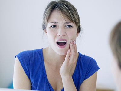 A woman with her mouth open in an expression of surprise or shock, looking at her reflection in a mirror while holding her hand up to her face.