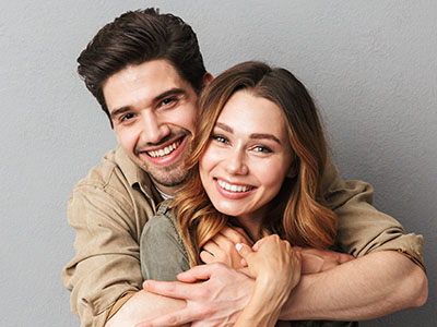 A man and woman embrace each other with smiles, both with short hair, standing close together against a neutral background.