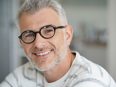 The image shows a man with glasses, smiling at the camera. He has gray hair and appears to be middle-aged.