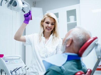 A dental hygienist is assisting an elderly man who is seated in a dentist s chair, both wearing face masks, with various dental equipment around them.