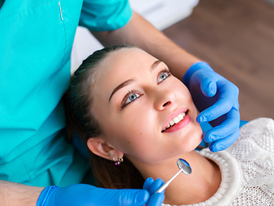 The image shows a person receiving dental care from a professional in a clinical setting, with the patient smiling at the camera while looking directly into it.
