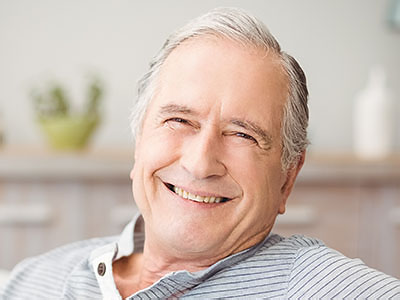 The image shows an elderly man with gray hair, wearing glasses, smiling broadly at the camera, sitting comfortably in a chair indoors.