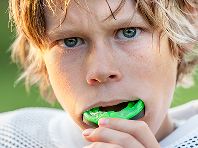 A young boy with blonde hair, wearing a white football jersey, brushing his teeth with a green toothbrush.