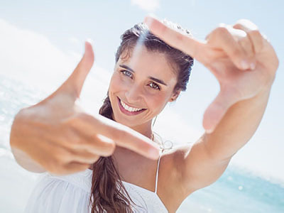 A woman with long hair is taking a selfie at the beach, smiling while holding her camera up to capture the moment.