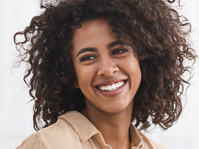 The image depicts a smiling woman with curly hair, wearing a white top and a beige blazer, standing against a light background.